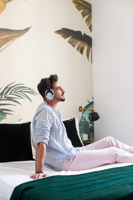 Бічний вид дбайливого бородатого іспанського чоловіка в навушниках сидячи на ліжку і насолоджуючись доброю музикою вдома. — стокове фото