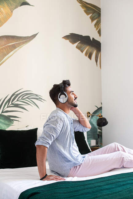 Бічний вид дбайливого бородатого іспанського чоловіка в навушниках сидячи на ліжку і насолоджуючись доброю музикою вдома. — стокове фото