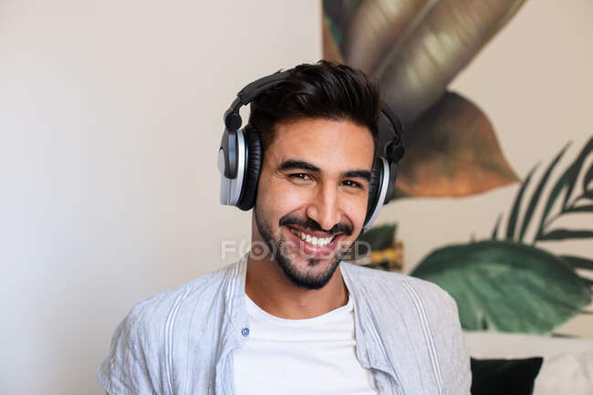 Heureux mec ethnique dans un casque souriant et regardant la caméra tout en écoutant de la musique à la maison — Photo de stock