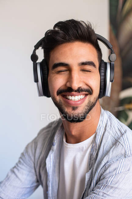 Heureux ethnique guy dans casque souriant avec les yeux fermés tout en écoutant de la musique à la maison — Photo de stock