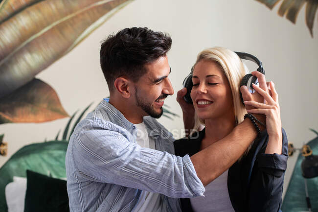 Fröhliche Frau mit Kopfhörern lächelt mit ihrem Freund, während sie zu Hause gemeinsam Musik hört — Stockfoto