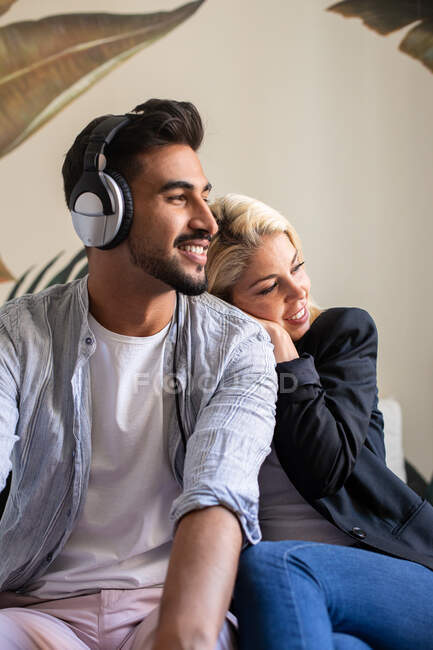 Веселая женщина, опираясь на плечо этнического бойфренда, слушает музыку в наушниках и смотрит в сторону, отдыхая дома вместе — стоковое фото
