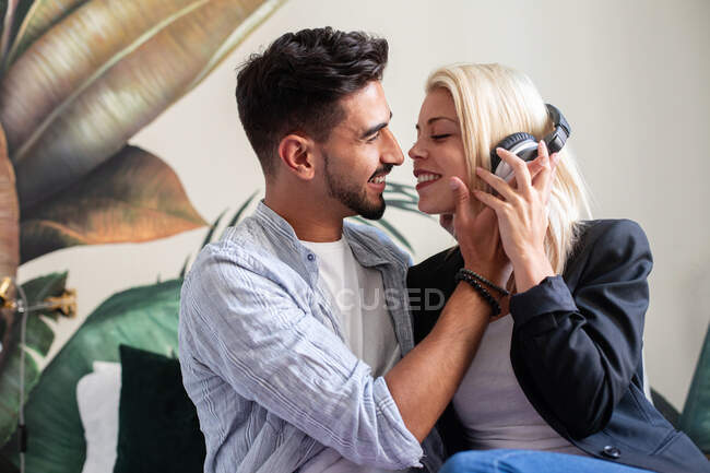 Веселая женщина в наушниках улыбается и пытается поцеловать этнического парня, слушая музыку дома вместе — стоковое фото