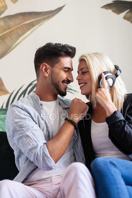 Donna allegra con le cuffie che sorride e cerca di baciare il fidanzato etnico ascoltando musica a casa insieme — Foto stock