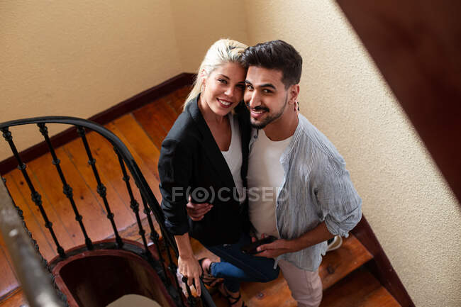 From above top view diverse man and woman smiling for camera and embracing each other while standing on stairway at home — Stock Photo
