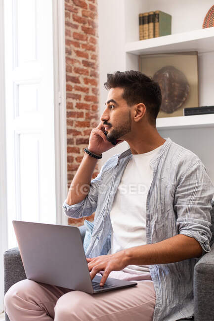 Bearded ethnic man using laptop and speaking on smartphone while working on remote project at home — Stock Photo