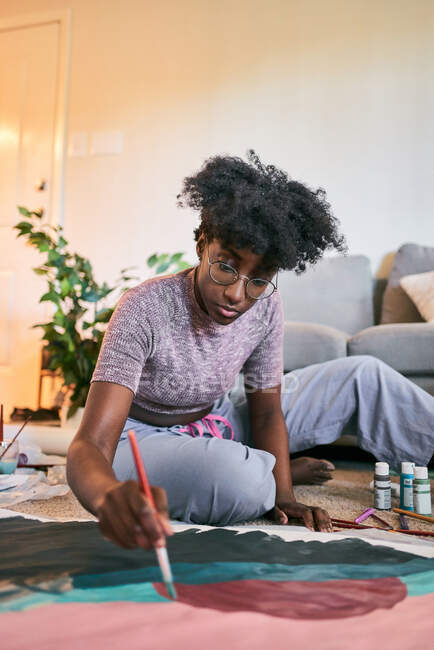Desde arriba de la cosecha anónima hembra negra con pincel dibujo colorido imagen en papel mientras está sentado en el suelo en casa - foto de stock