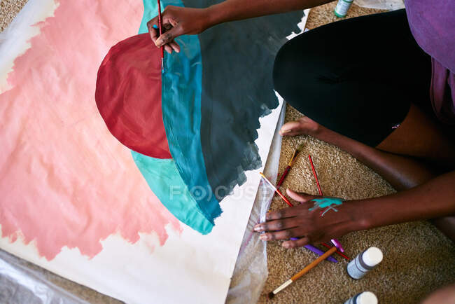 Dall'alto del raccolto anonimo femmina nera con pennello disegno colorato immagine su carta mentre seduto sul pavimento a casa — Foto stock