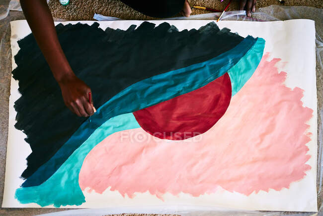 Сверху анонимная черная женщина с кистью, рисующая красочную картину на бумаге, сидя дома на полу — стоковое фото