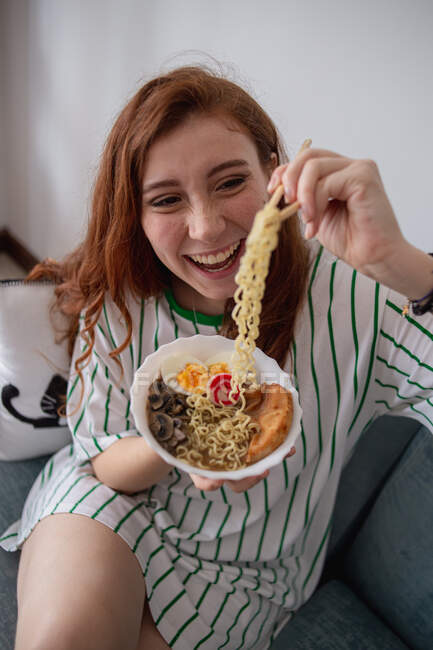 From above cheerful redhead female laughing and picking noodles from bowl of tasty ramen while sitting on couch at home — Stock Photo