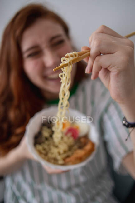 Blurred cheerful redhead female laughing and picking noodles from bowl of tasty ramen while sitting on couch at home — Stock Photo