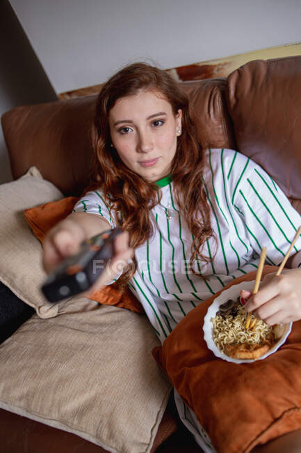 From above young redhead woman eating ramen and changing channels on TV while sitting on sofa during lunch at home — Stock Photo