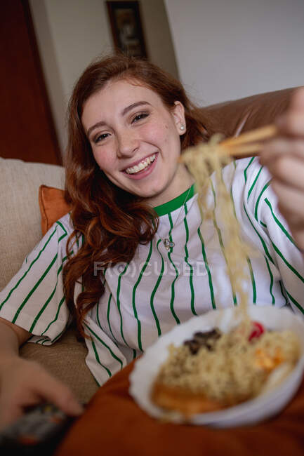 D'en haut jeune rousse femme manger ramen et changer de chaînes à la télévision tout en étant assis sur le canapé pendant le déjeuner à la maison — Photo de stock