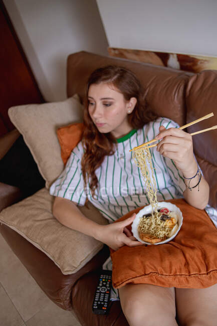 De cima jovem ruiva mulher comendo ramen e mudando de canais na TV enquanto sentado no sofá durante o almoço em casa — Fotografia de Stock