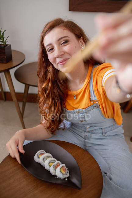 Сверху молодая рыжая женщина в повседневной одежде смотрит в камеру палочками, сидя за столом и поедая суши дома. — стоковое фото