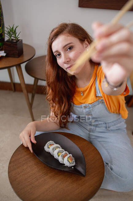 D'en haut jeune rousse femme en vêtements décontractés regardant la caméra à l'aide de baguettes tout en étant assis à la table et en mangeant des sushis à la maison — Photo de stock