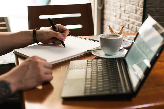 Homme méconnaissable esquissant dans un bloc-notes près d'une tasse de café et d'un ordinateur portable alors qu'il était assis à table et travaillait à un projet à distance à la cafétéria — Photo de stock