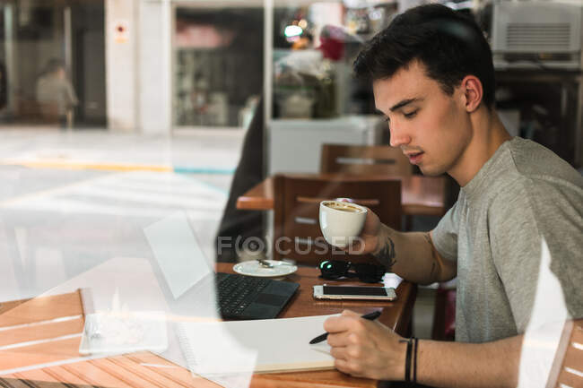 Vista lateral del joven con taza de café escribiendo en bloc de notas mientras está sentado en la mesa detrás del vidrio y estudiando en la cafetería - foto de stock