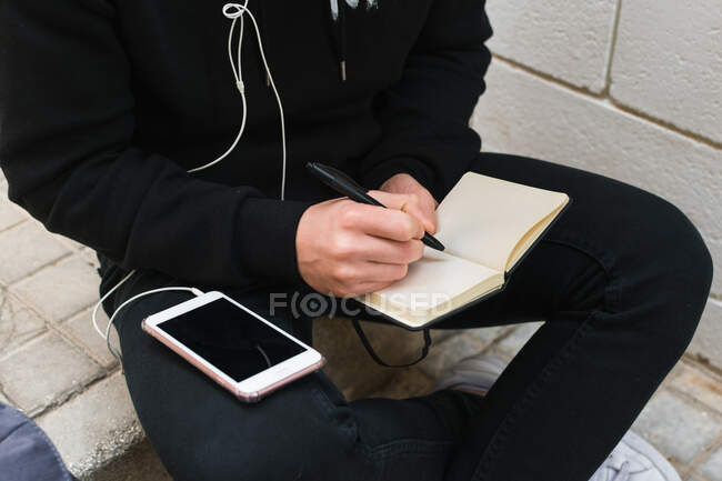 Сверху анонимный парень в повседневной одежде слушает музыку на смартфоне и делает заметки в блокноте, сидя на ступеньках на городской улице — стоковое фото