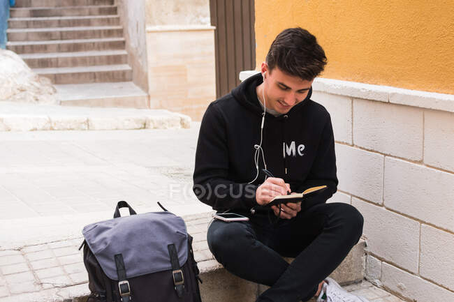 Cheerful young man in casual clothes listening to music in earphones and writing in notebook while sitting on step near backpack on city street — Stock Photo