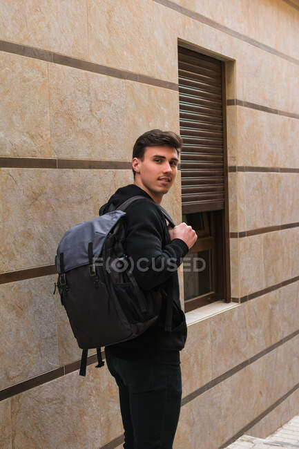 Side view of positive young man with backpack looking at camera while standing near window of modern building on city street — Stock Photo