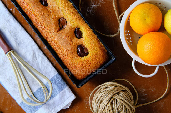 Vista dall'alto di pan di Spagna appena sfornato torta al cioccolato e arance mature poste sul tavolo vicino frusta e filo in cucina — Foto stock
