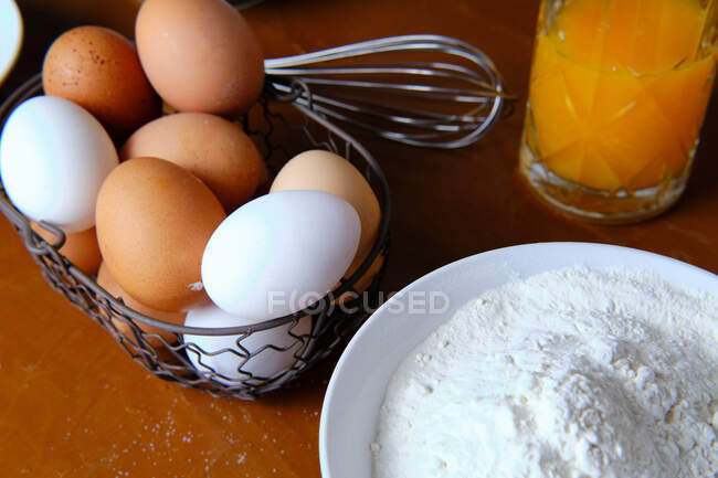 Glass of fresh citrus juice on table near bowl with flour and basket of raw eggs during pastry preparation at home — Stock Photo