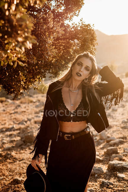 Похмура жінка в стильному чорному сільському одязі торкається волосся і дивиться на камеру, стоячи під гілками дерев під час заходу сонця в сільській місцевості. — стокове фото