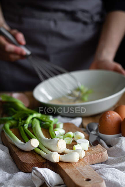 Ein Bündel frischer Schalotten und Pilze auf einem Schneidebrett in der Nähe von Eiern und Mohn gegen die Hausfrau, die Zutaten in einer Schüssel vermischt — Stockfoto