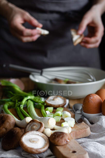 Ein Bündel frischer Schalotten und Pilze auf einem Schneidebrett in der Nähe von Eiern und Mohn gegen die Hausfrau, die Zutaten in einer Schüssel vermischt — Stockfoto