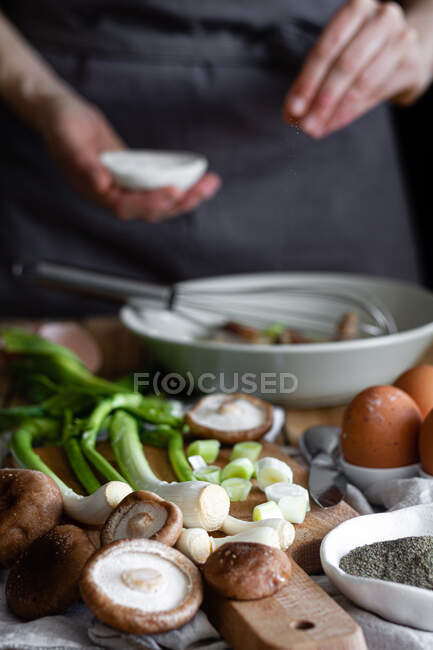 Bando de cebolinhas frescas e cogumelos colocados na tábua de corte perto de ovos e sementes de papoula contra a dona de casa da colheita misturando ingredientes na tigela — Fotografia de Stock