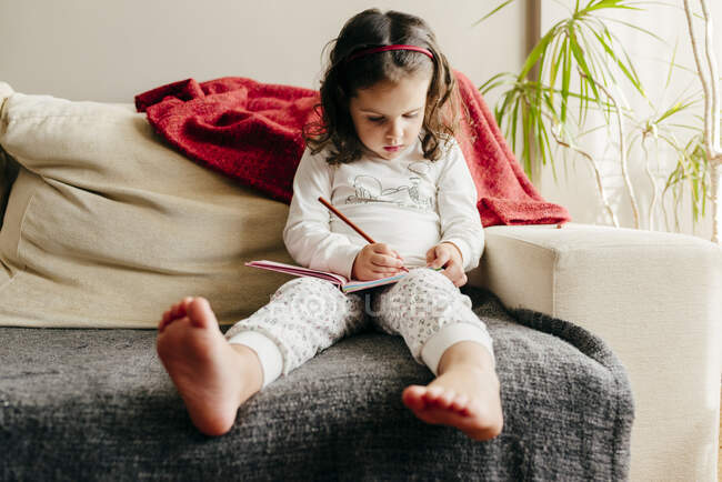 Liebenswertes barfüßiges Mädchen im Pyjama sitzt auf der Couch und zeichnet im Skizzenbuch im gemütlichen Wohnzimmer zu Hause — Stockfoto