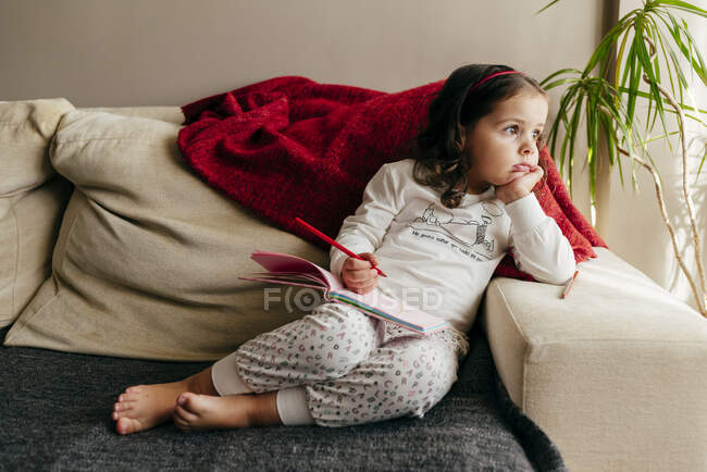 Jolie petite fille assise sur le canapé avec un cahier à la recherche d'inspiration — Photo de stock