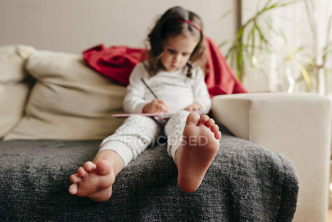 Petite fille pieds nus non concentrée assise sur le canapé et dessinant dans le carnet — Photo de stock