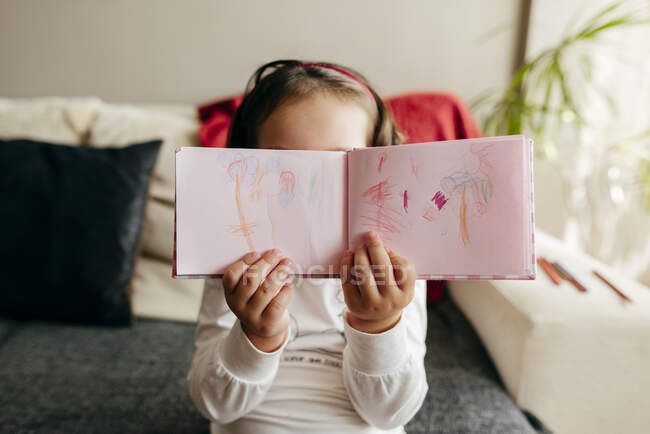 Неузнаваемая девочка демонстрирует тетрадь с рисунками, сидя на диване и рисуя дома — стоковое фото
