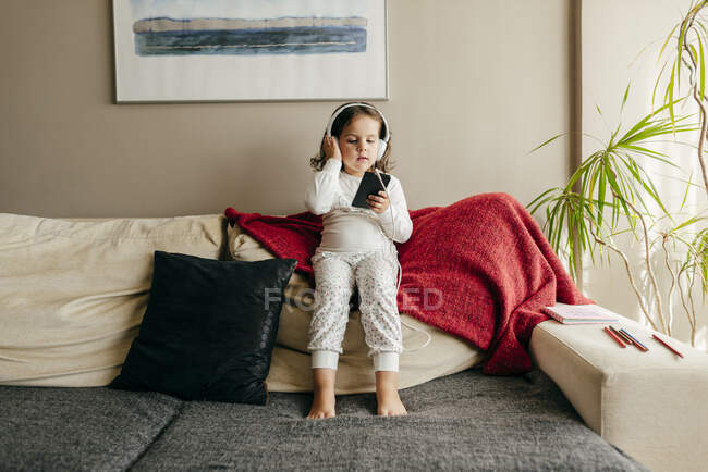 Carino bambina seduta sul divano ad ascoltare musica con le cuffie — Foto stock