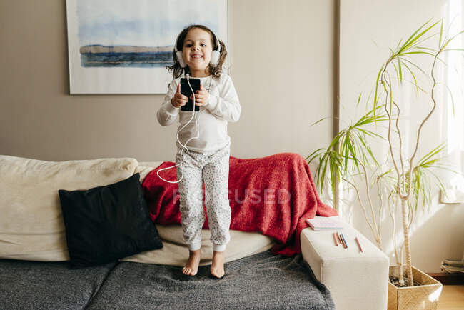 Petite fille mignonne sautant sur le canapé et écoutant de la musique avec des écouteurs — Photo de stock