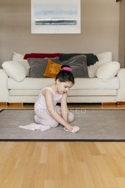 Vista lateral da menina bonito em collants e collants sentado no chão perto do sofá e vestindo sapatos de dança antes do ensaio de balé em casa — Fotografia de Stock