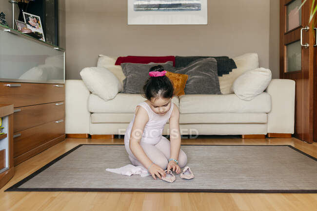Cute girl in leotard and tights sitting on floor near sofa and putting on dance shoes before ballet rehearsal at home — Stock Photo