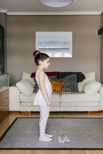 Nettes kleines Mädchen in hellrosa Trikot und Strumpfhosen beim Stehen neben Tanzschuhen im gemütlichen Wohnzimmer zu Hause — Stockfoto