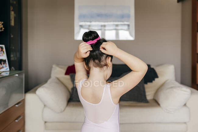 Vista posterior de la pequeña bailarina anónima linda en maillot haciendo bollo de pelo antes del entrenamiento de ballet en la acogedora sala de estar en casa - foto de stock