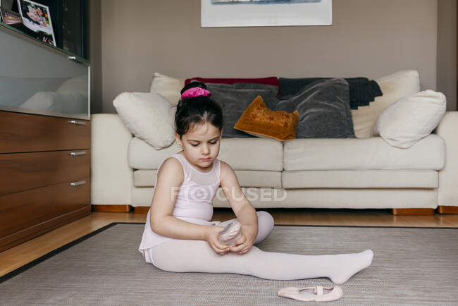 Seitenansicht des süßen Mädchens in Trikot und Strumpfhose, das auf dem Boden neben dem Sofa sitzt und vor der Ballettprobe zu Hause Tanzschuhe anzieht — Stockfoto