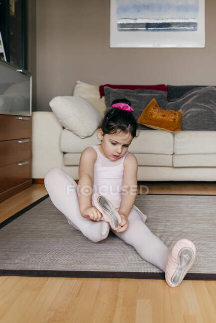 Милая девушка в трико и трико сидит на полу рядом с диваном и надевает танцевальную обувь перед репетицией балета дома — стоковое фото