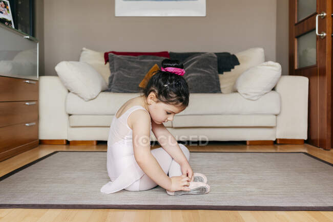 Vista laterale della ragazza carina in body e collant seduti sul pavimento vicino al divano e indossando scarpe da ballo prima delle prove di balletto a casa — Foto stock