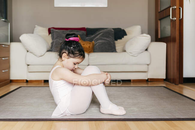 Скучная задумчивая грустная маленькая девочка в трико и трико, сидящая на полу и отдыхающая во время репетиции балета дома — стоковое фото