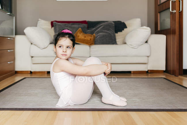 Скучная задумчивая маленькая девочка в трико и трико, сидящая на полу и смотрящая вдаль во время репетиции балета дома — стоковое фото