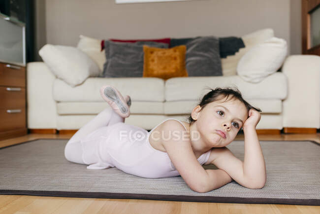 Bored thoughtful little girl in leotard lying on the floor looking away while resting during ballet rehearsal at home — Stock Photo