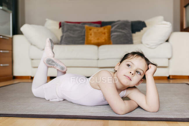 Скучная задумчивая маленькая девочка в трико, лежащая на полу и смотрящая в сторону во время репетиции балета дома — стоковое фото