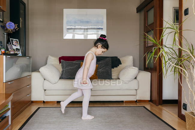 Vista lateral de la linda niña mirando hacia otro lado bailando cerca del sofá durante el ensayo de ballet en casa - foto de stock