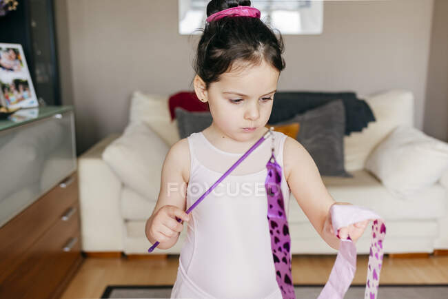 Орієнтована мила дівчинка брюнетка в леопарді, дивлячись геть, обертаючись стрічкою під час тренування ритмічної гімнастики в затишній вітальні вдома — стокове фото
